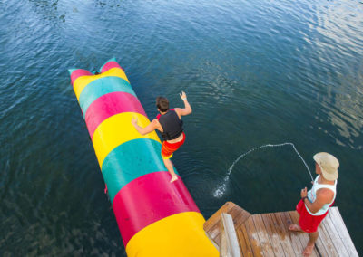 summer staff lifeguard watches man jump off deck onto inflatable pillow on lake