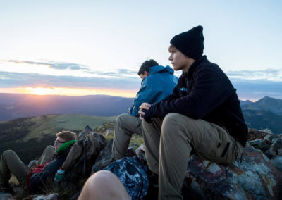 three guys sit on rocks at mountain summit overlooking sunset